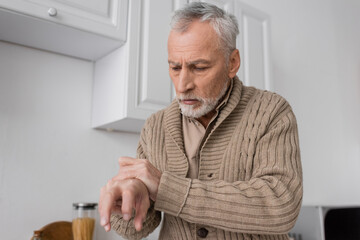 grey haired man with parkinson disease looking at trembling hands in kitchen.