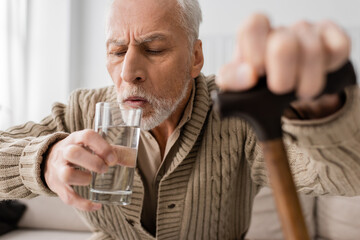 grey haired man with parkinson disease and trembling hands drinking water while sitting with...