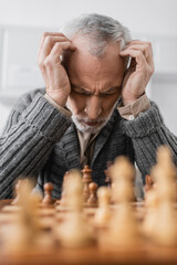 depressed man with alzheimer disease sitting with closed eyes and hands near head at chessboard on blurred foreground.