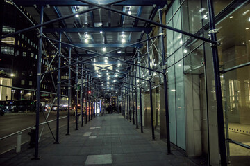 View down an empty city sidewalk at night with metal scaffolding and hoarding and lights, nobody