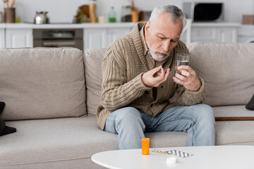 senior man with parkinsonian syndrome holding pill and glass of water while sitting on couch near medication on table.