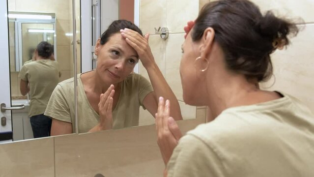 Age changes on the face. A cute adult woman looks at her face in the mirror in the bathroom.