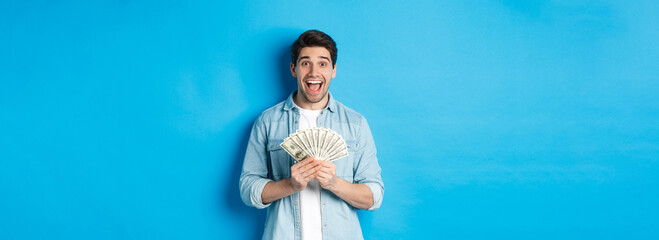 Excited and surprised attractive man, holding money prize and smiling amazed, standing over blue...