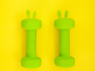 two green heavy dumbbells with leaves in shape of Easter bunny on yellow background. Easter fitness and training composition with copy space. Top view from above, flat lay