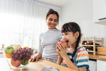 Caucasian little kid biting an apple with mother in kitchen room. 