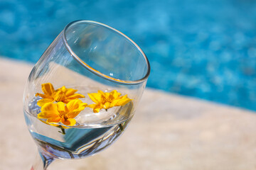 Glass of water with flowers near swimming pool, closeup