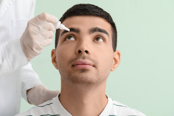 Ophthalmologist putting drops in young man's eye on green background, closeup