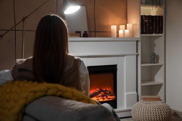 Young woman sitting on sofa near fireplace at home in evening, back view