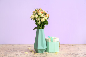 Vase with beautiful rose flowers and gift box on table against color background