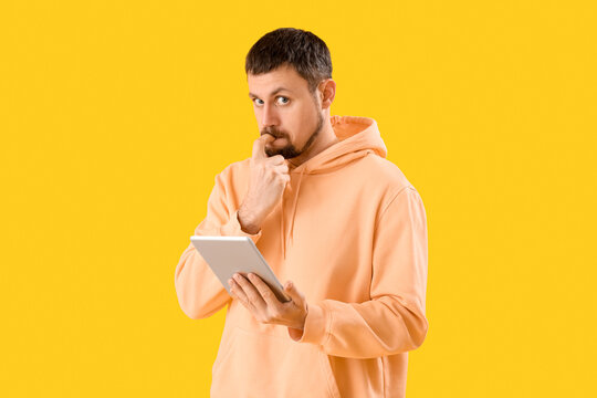 Handsome man with tablet computer biting nails on yellow background
