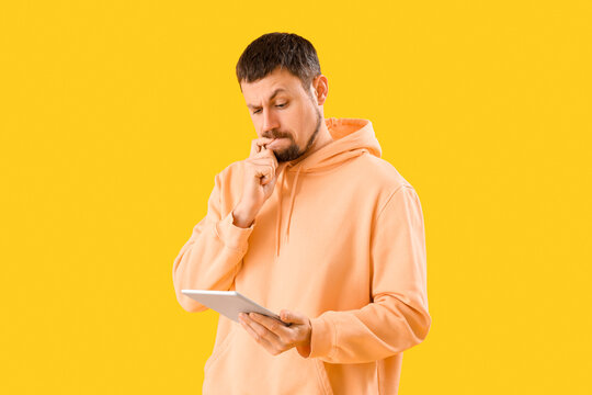 Handsome man with tablet computer biting nails on yellow background