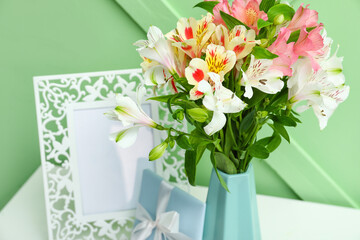 Vase with beautiful alstroemeria flowers, gift and picture frame on table near color wall, closeup. Mother's day celebration