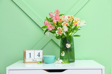 Vase with beautiful alstroemeria flowers, cup of coffee and cube calendar on table near color wall.  Mother's day celebration
