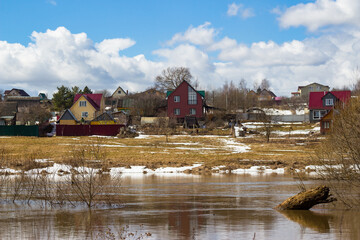 Rural buildings on a hill beyond the river against a blue sky with clouds, a river overflowing in...