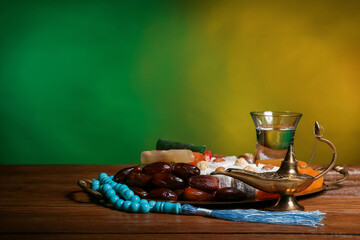 Aladdin lamp of wishes, prayer beads, glass and sweets for Ramadan on table against color background