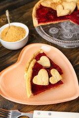 Heart-shaped plate with pieces of sweet strawberry pie, cookie hearts and bowl of brown sugar on wooden table