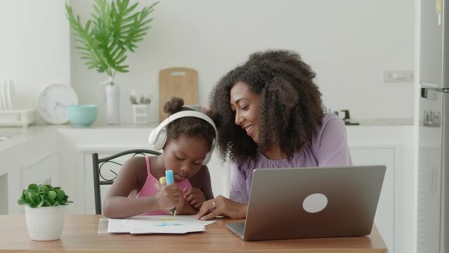 Happy multiethnic women single mom sits next adorable baby girl in wireless headphones Mom her baby girl drawing together colorful images with felt-tip pens watching online course on laptop at home