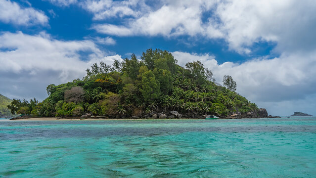 The beautiful tropical island is completely overgrown with lush vegetation - palm trees, bushes. Boulders piled up near the shore. The boat is moored in the turquoise ocean. Blue sky, clouds. 