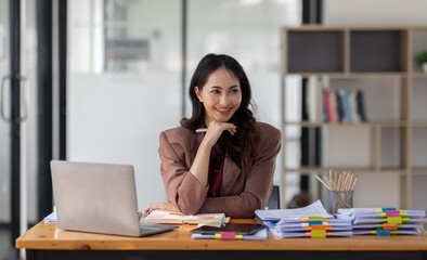 Freelancer Happy business Asian woman in knitwear taking notes at laptop sitting at desk office, finance concept.
