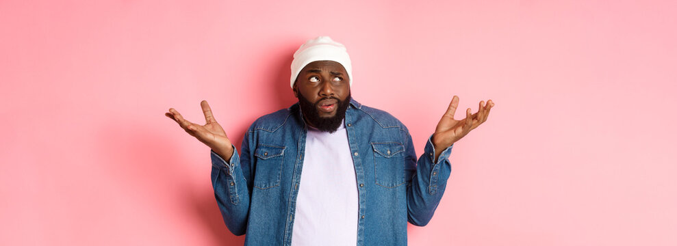 Confused African-american Man In Beanie Looking At Upper Left Corner Doubtful, Shrugging Uncertain, Standing Over Pink Background