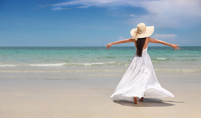 Fototapeta na wymiar Back view young happy asian traveller woman raising hands with beautiful dress and hat standing on sandy beach. Cute girl enjoy tropical sea on relax holiday vacation during summer and sunshine day