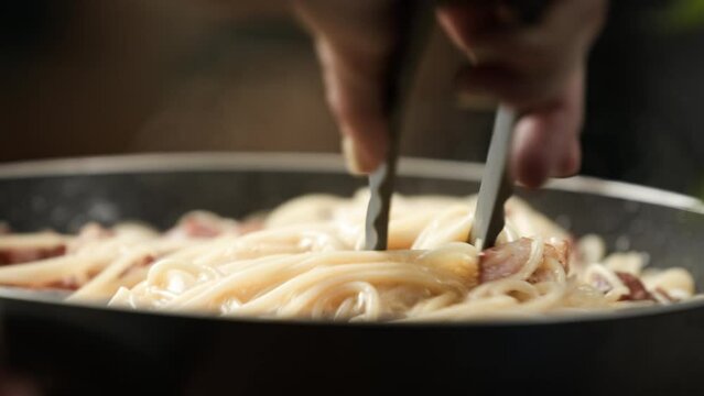 Carbonara recipe; stirring cooked spaghetti pasta with beaten eggs and bacon using tongs; creamy thick texture; close up, 4k, slow motion