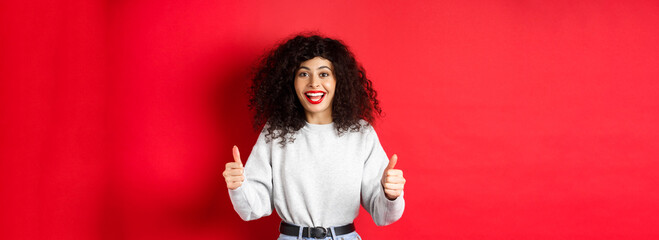 Enthusiastic girl with curly hair and red lips, showing thumbs up and saying yes, agree with you,...