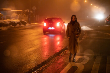 a young woman in a long coat stands by the road with passing cars at night rainy autumn city