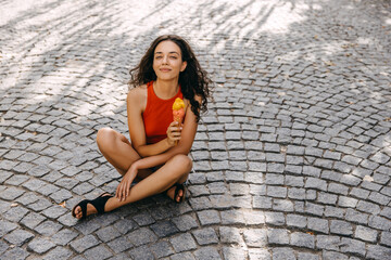 Young brunette woman sitting on city street on a hot summer day, holding an ice cream and smiling.