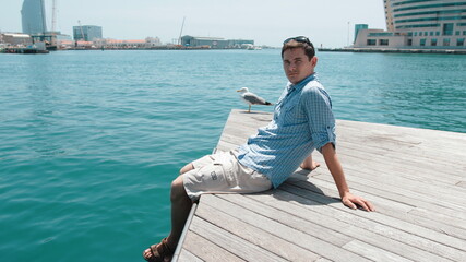 A guy in a blue shirt sits on a pier next to a seagull