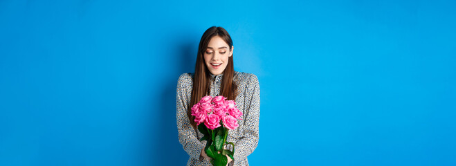 Valentines day concept. Happy attractive woman receive surprise flowers, looking thankful at bouquet of pink roses, standing on blue background