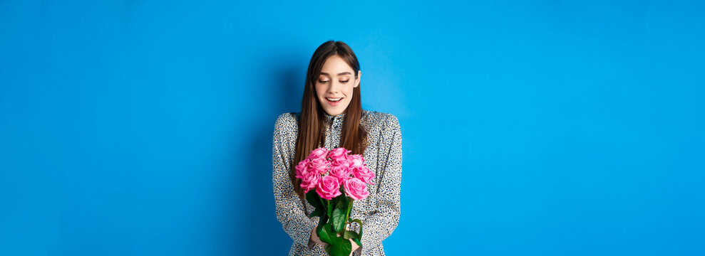 Valentines day concept. Happy attractive woman receive surprise flowers, looking thankful at bouquet of pink roses, standing on blue background
