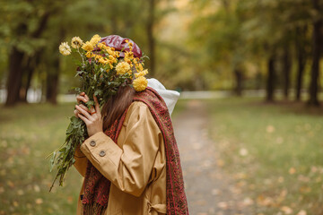 Side view of unrecognizable shy woman covering face with floral bouquet of yellow blooms in autumn...