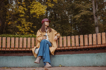 Full length of relaxed woman in trench sitting on bench with takeaway coffee cup in autumn park 