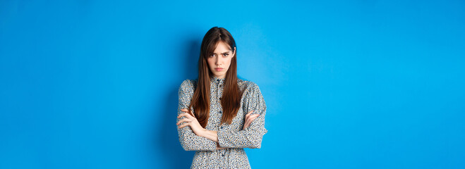 Angry young woman in dress feel unfair, cross arms on chest and frowning offended, standing on blue...