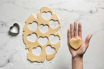 A heart shaped shortbread raw cookie on a palm over rolled out cookie dough, baking at home - 562650008