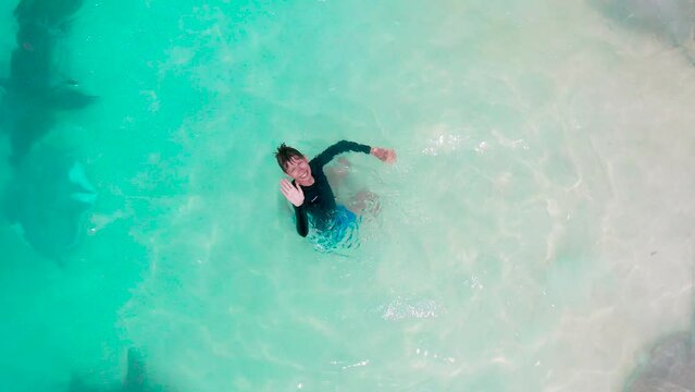 A young boy swims among big nurse sharks and stingrays in the turquoise sea with white sand beach of Fulidhoo Island in the Maldives. Aerial top view.