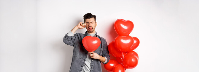 Valentines day and love concept. Sad crying man holding red heart balloon and whiping tears,...