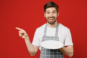 Young waiter male housewife housekeeper chef cook baker man wear grey apron hold empty plate point...