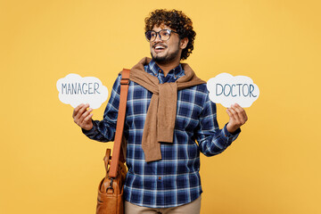 Young smiling teen Indian boy student wears casual clothes glasses bag near card with doctor manager text choose profession look aside on area isolated on plain yellow background University concept.