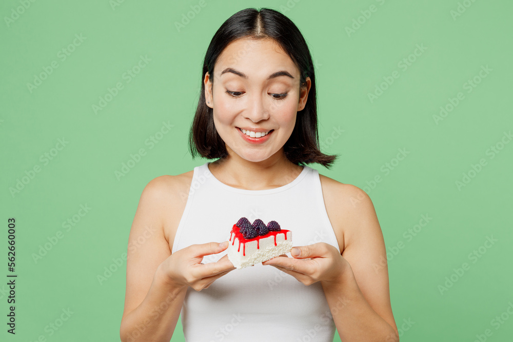 Wall mural Young smiling happy fun woman wearing white clothes holding in hand pice of cake dessert isolated on plain pastel light green background. Proper nutrition healthy fast food unhealthy choice concept.