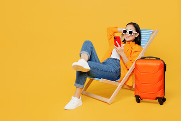 Young fun woman wear summer clothes sit in deckchair use mobile cell phone isolated on plain yellow background. Tourist travel abroad in free spare time rest getaway. Air flight trip journey concept. - obrazy, fototapety, plakaty