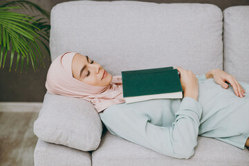 Young muslim woman wearing hijab casual clothes read book sleeping lying on sofa couch stay at home flat rest relax spend free spare time in living room indoor People middle eastern uae islam concept