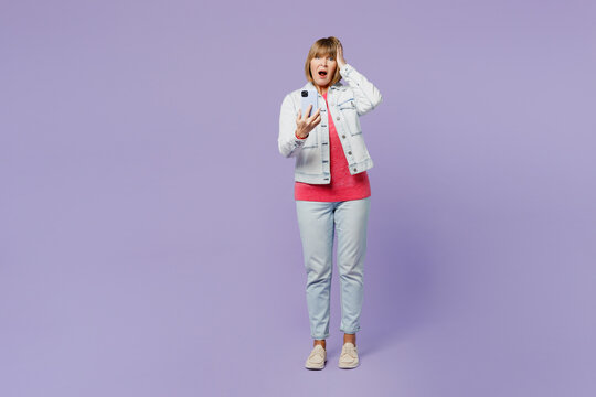 Full body shocked sad elderly blonde woman 50s years old she wear casual clothes denim jacket t-shirt hold head use mobile cell phone isolated on plain pastel light purple background studio portrait.