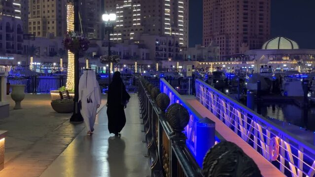 Arab couple man with traditional white long tidy clean dress and woman with black long dress Islamic hijab are walking at night in a luxury scenic wonderful city landscape in Pearl Doha Qatar island