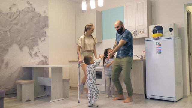 A Family Of Four Mom Dad Daughter And Son Have Fun And Dance In The Kitchen At Home.A Young Beautiful Caucasian Girl Is Having Fun And Dancing On One Crutch.Happy Family, Children In Dad's Arms