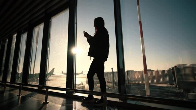 Silhouette young female traveler at airport, holding smart phone device, at background planes outdoors, airport runway. Using wireless mobile technology. Rest in tour at travel. Business trip.