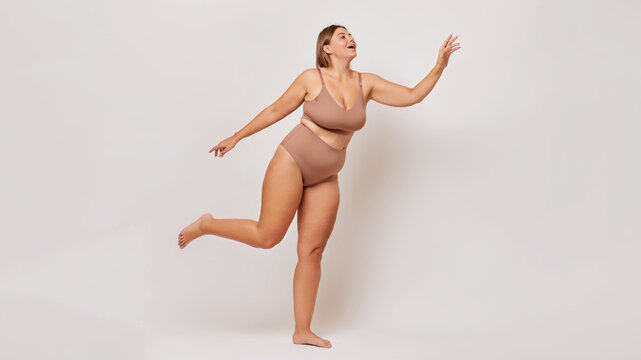 Body positive concept. Charming young woman wears basic lingerie flies in air and smiles happily feels confident, poses over white background