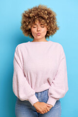 Vertical shot of cute young woman with curly Afro hair keeps eyes closed lips folded waits for kiss, dreams about something, wears pink pullover, isolated over blue wall