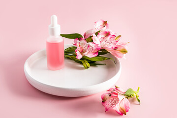 A beautiful cosmetic bottle with a white pipette stands on a ceramic plate with astromeria flowers. pink background. natural cosmetics.